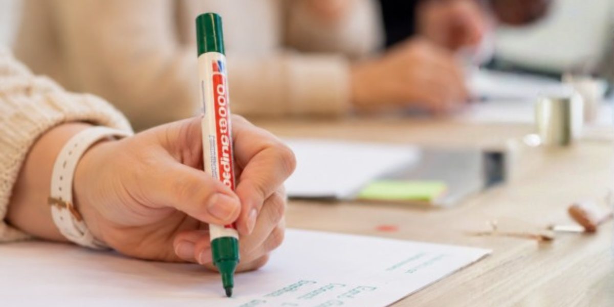 Close-up van een hand die met een groene stift op papier schrijft tijdens een gezamenlijke werksessie.