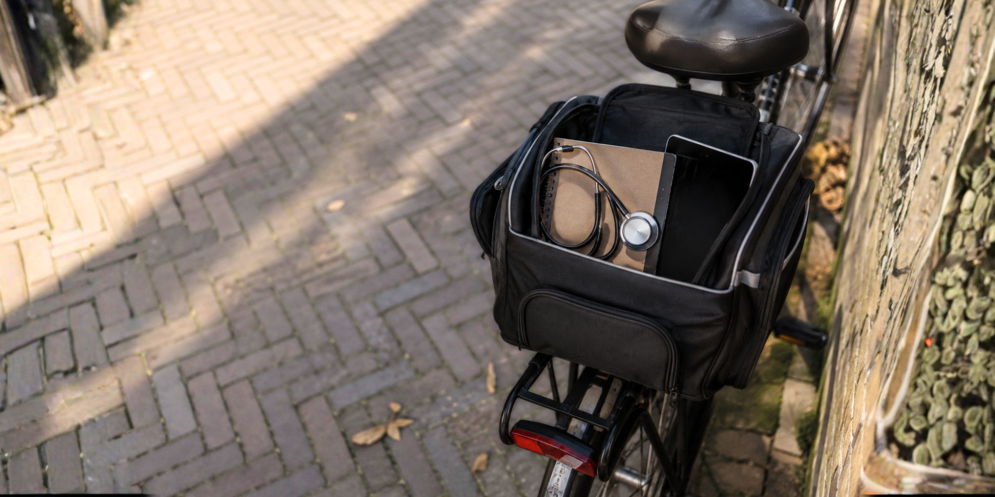 Een zwarte fietstas achter op een fiets met daarin een stethoscoop, notitieboek en tablet, klaar voor een huisbezoek door een zorgprofessional.